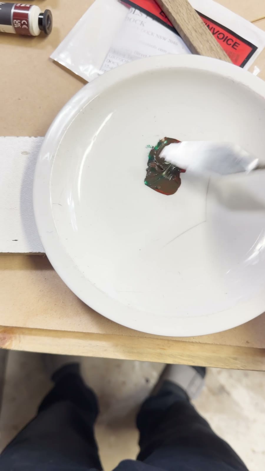 Green and brown paint being mixed with a palette knife on a white plate at the workbench.